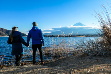 Fototapeta na wymiar A couple holding hands at the side of Kawaguchiko Lake, Japan with the view on Mt Fuji. The mountain surrounded by clouds, top of it covered with snow. Exploring new places. Love and affection