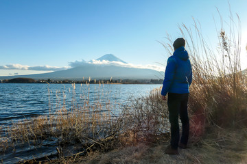 Fototapeta na wymiar A man walking in between golden grass at the shore of Kawaguchiko Lake, Japan with the view on Mt Fuji. The man is enjoying the view on the volcano. The mountain surrounded by clouds. Serenity