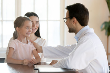 Smiling male doctor stroking small cute girl patient, encouraging before checkup. Young mommy holding on lap happy little daughter, getting acquainted with pediatrician at meeting in hospital.