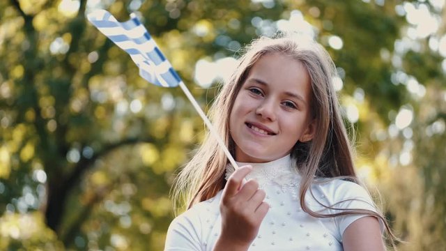 Girl holds Greek flag. The background is blurred. Kid is in park with white and blue flag.