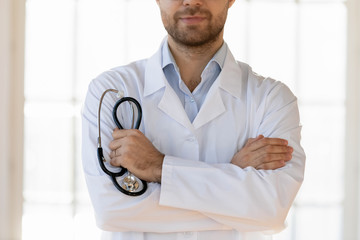 Crop close up confident male general practitioner standing with folded hands, holding stethoscope....