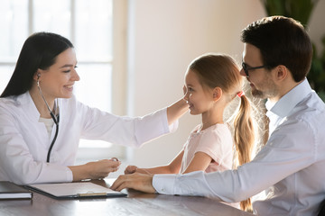 Obraz premium Smiling pleasant female pediatrician stroking head of preschool girl sitting on father s lap. Smiling doctor getting acquainted with small patient before starting checkup listening heartbeat lungs.