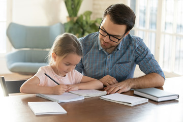 Fototapeta na wymiar Caring young father helping little daughter with homework. Attentive small schoolgirl studying at home with young daddy. Dad teaching child writing letters in copy book, children education concept.