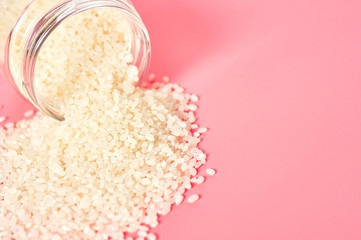 White rice poured out of glass jar on pink background. Copy space