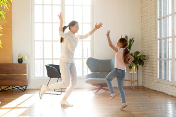 Obraz premium Full length excited young mommy repeating dance moves for school performance with happy small daughter. Joyful little preschool kid girl having fun together with cheerful mother in living room.