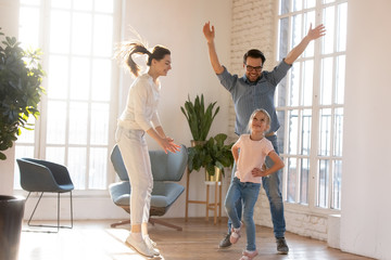 Overjoyed young married couple having fun, watching little preschool daughter dancing in living...