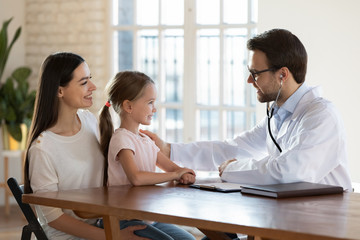 Side view friendly male doctor holding hand on little cute patient shoulder. Smiling young mother holding on lap small daughter, listening to doc conclusion after checkup examining health condition.