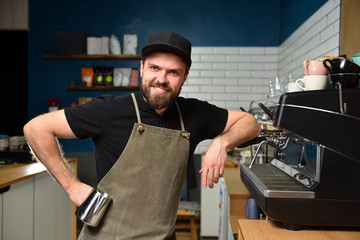 Young bearded barista guy smiles at work in a coffee shop leaning on a coffee machine