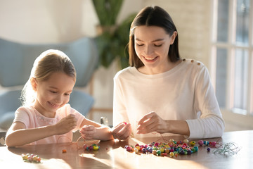 Happy small preschool kid girl sitting at table with elderly sister, creating handmade bracelets with wooden supplies at home. Smiling young mom nanny stringing beads on needles with little daughter.