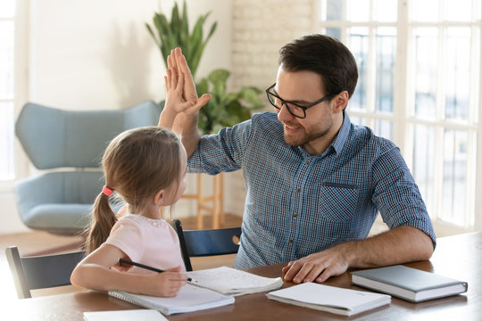 Happy young dad giving high five to smiling little daughter, satisfied with homework. Joyful male tutor praising small girl with successful exercise finish. Home schooling children education concept.
