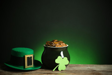 Pot with gold coins, hat and clover on wooden table against dark background, space for text. St....