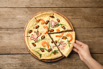 Woman taking piece of delicious seafood pizza at wooden table, top view