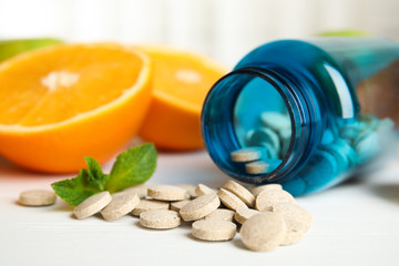 Bottle with vitamin pills and orange on white wooden table, closeup