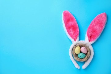 Headband with Easter bunny ears and dyed eggs in nest on light blue background, flat lay. Space for text