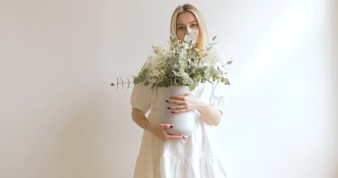 Blonde Woman In A White Dress With Flowers In Hands Is Posing On The Background