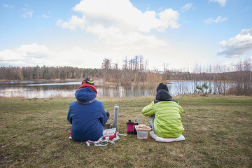 Kids performing picnic outdoors near lake in early spring, view from back