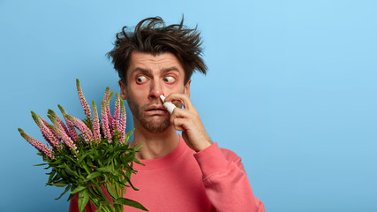 Unhappy man drips nose, looks in dissatisfiaction at sourse of allergy, has sick tired look, suffers from rhinitis, wears pink jumper, isolated on blue background. Place for your advertising - obrazy, fototapety, plakaty