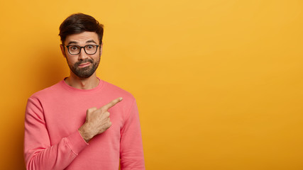 Surprised cheerful unshaven man advices special discount offer, points at upper right corner to advertise product, poses against yellow background, dressed casually, shows promo, invites you to party