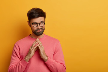 Fototapeta premium Tricky thoughtful bearded man steepls fingers, has some intention in mind, considers plans for future, looks aside mysteriously, has scheme, wears rosy sweater, isolated over yellow background