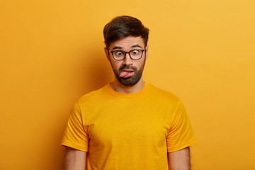Portrait of bearded guy shows grimace, crosses eyes and sticks out tongue, plays around, going crazy, wears spectacles, everyday t shirt, poses against yellow background. Human face expressions