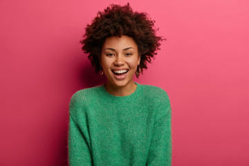 Portrait of good looking curly ethnic woman smiles broadly, enjoys day off, has happy talk with interlocutor, discusses holiday preparation, wears green sweater, isolated on pink background.