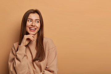 Pleasant looking cheerful brunette woman looks aside with charming smile, keeps hand under chin, wears casual sweatshirt, poses against beige background, remembers hilarious event, has joyful look