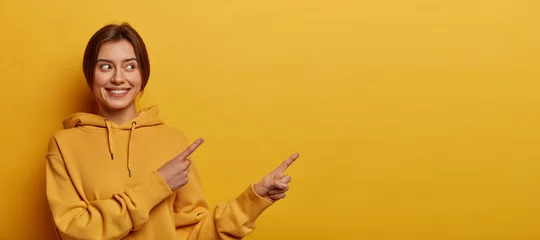 Foto op Canvas Smiling dark haired woman gives advice and points right, promots product, wears casual sweatshirt, poses against yellow background, indicates at advertisement, introduces promo with pleased expression © Wayhome Studio