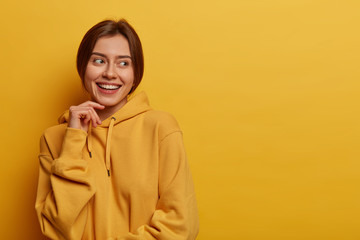 Pleasant looking Caucasian woman keeps hand on face, looks away with satisfaction, notices...