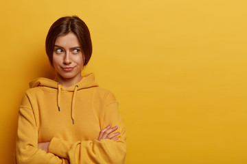 Fototapeta premium Horizontal shot of serious looking European woman crosses hands over chest, thinks about something with doubt, wears casual hoodie, poses against yellow studio wall, doesnt want to talk to you