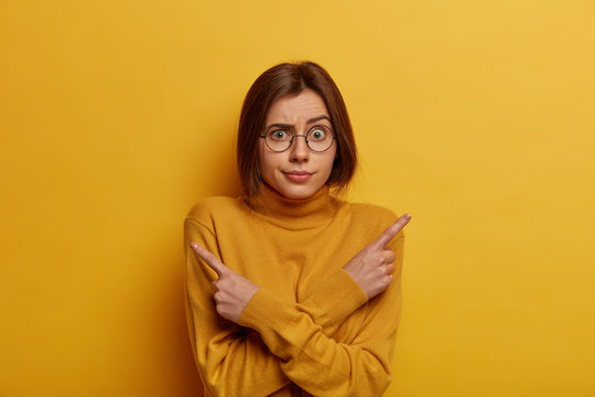 Studio shot of embarrassed unaware woman points sideways, thinks what to choose, raises eyebrows, indicates right and left, dressed in yellow outfit, stands indecisive indoor. Difficult choice