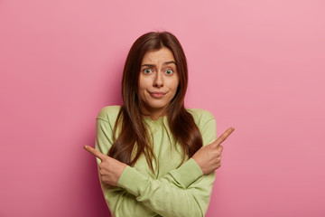 Confused young woman with dark hair indicates sideways, faces opportunity to choose, has hesitant expression, wears green jumper, isolated over pink pastel backgrounnd. What to select I wonder