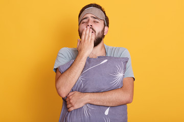 Horizontal shot of handsome tired brunet man wearing gray t shirt, embracing grey pillow isolated over yellow background. Resting after long day. Bearded guy with closed eyes and sleeping mask.