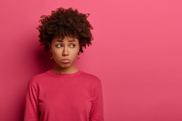 Obraz na płótnie Canvas Disappointed sad woman with Afro hair looks unhappily aside, being bothered by something, regrets because of prohibition, wears rosy jumper in one tone with background. Unhappy feelings concept