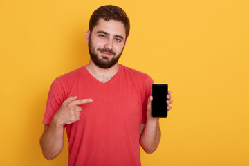 Indoor shot of man holding smartphone in his hands and pointing at black screen with his index finger isolated over yellow background. Bearded man wearing red t shirt and showing phone. Copy space.