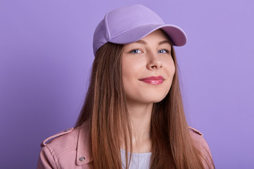 Fototapeta premium Picture of beautiful young pleasant blonde girl, lady wearing cap and jacket, fashionable girl looking directly at camera, posing isolated over lilac studio background, looks happy and satisfied.