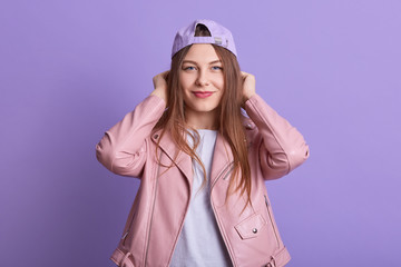 Close up portrait of pretty young woman wearing cap, white shirt and pink leather jacket, attarctive female posing isoalted over lilac studio background with hands on her head and looking at camera.