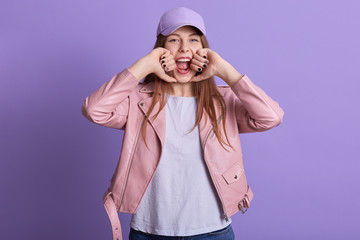 Horizontal shot of woman with long fair hair poses in studio dressed white shirt, pink leather jacket and cap, attractive young female screaming something, keeps fists near her face against lilac wall