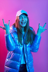 Image of laughing good looking cute young female opening mouth widely, wearing jacket and cap, raising arms, making gesture, showing sign of horns, being in high spirits. Neon lights concept.