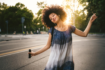 Young woman dancing listening to the music with earphones from the smart phone outside a park at sunset on a summer day
