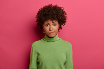 Fototapeta na wymiar Puzzled concerned female purses lips and looks nervously at camera, has worried expression, wears green turtleneck, isolated on pink background, ponders on right decision. Human reaction concept