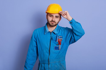 Portrait of serious responsible young worker holding his yellow helmet with one hand, wearing uniform, having equipment in pockets, standing isolated over blue background in studio. Work concept.