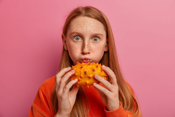Horizontal shot of lovely freckled child poses with exotic fruit, blows cheeks and stares with bugged eyes, holds ripe fresh horned melon, has long ginger hair, models agaist pink studio background.