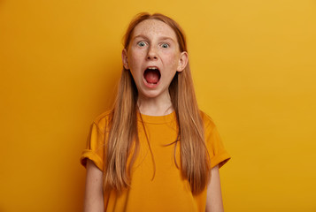 Emotive small girl keeps mouth widely opened, screams loudly, has freckled skin, wears yellow t shirt, watches horror film, poses indoor. Female teenager exclaims from wonder, feels excited.