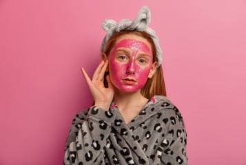 Portrait of cute tender foxy child applies beauty mask, cares about face, has healthy fresh skin at home, looks direclty at camera, isolated over pink background. Children, cosmetology concept