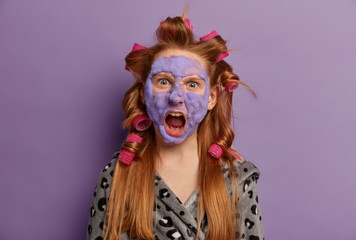 Isolated shot of angry emotional redhead teenage girl yells loudly, keeps mouth widely opened, wears hair curlers and cosmetic mask, maintains beauty procedures, poses over purple background