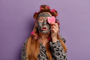 Shocked young female model applies clay mask, covers eye with cosmetic sponge, has beauty treatments, wears hair curlers, prepares for party, models indoor. Children, beauty, wellness concept