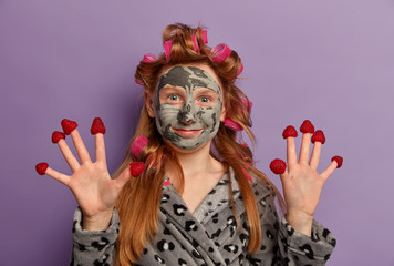 Happy teenage girl applies facial clay mask, wears hair curlers, raises palms, has raspberry on fingers, enjoys beauty procedures, models against purple background. Children, cosmetology, spa
