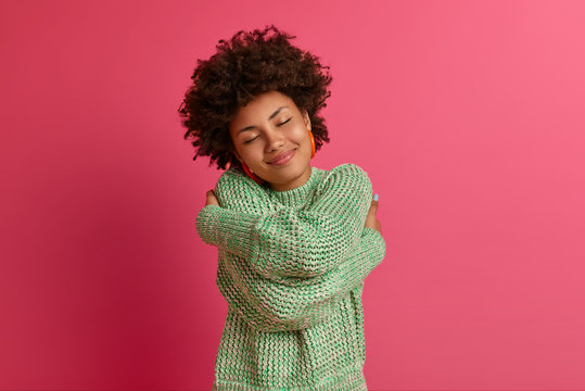 Tender curly Afro American woman cuddles herself, closes eyes and tilts head, feels coziness, wears warm sweater, dreams about something pleasant, poses against rosy background. Self love concept