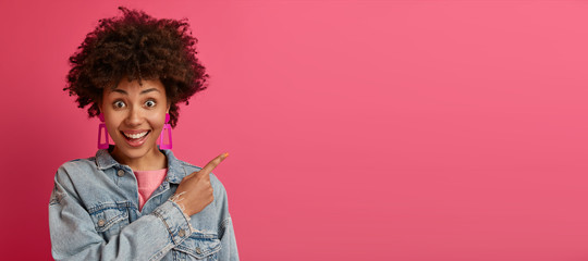 Joyful Afro American woman smiles broadly, gives advice where to find what you need, promots product, recommends advertisement, discusses new store, wears jean jacket, isolated over pink background