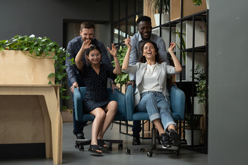Diverse colleagues having fun, riding on chairs in modern office, excited employees laughing, enjoying funny game during break, celebrating business success or achievement, team building activity